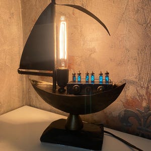 Może przedstawiać: Lampa biurkowa w stylu steampunk w kształcie żaglówki. Lampa ma ciemne metalowe wykończenie, żarówkę w stylu Edisona i cyfrowy wyświetlacz zegara. Podstawa to ciemny drewniany blok. Lampa jest podłączona do gniazdka elektrycznego.