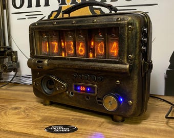 Può includere: Un orologio a tubi nixie rettangolare, metallico e color bronzo con una maniglia superiore. Il dispositivo presenta sei tubi nixie arancioni luminosi che mostrano l'ora, un piccolo display digitale blu, un altoparlante e varie manopole.