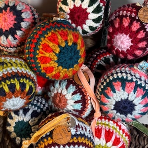 May include: A collection of spherical ornaments covered in multi-colored crochet patterns. Each ornament features a circular design with radiating colors including red, green, blue, yellow, and pink. Small wooden tags labeled pure joy are attached.
