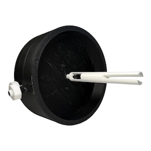 May include: A black cylindrical spool holder featuring a textured interior surface and a central white plastic spindle with a split-end design. A small white nut and a black screw are attached to the exterior side of the black circular housing.