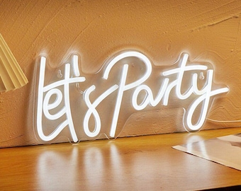 May include: A white neon sign that reads "Let's Party" in a cursive font. The sign is illuminated and sits on a wooden surface, with a textured, tan wall in the background. The sign is a decorative item.