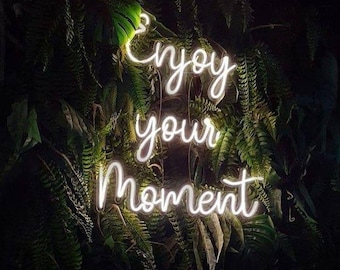May include: A neon sign with the words "Enjoy your Moment" in a cursive font. The sign is illuminated in a bright white color and is set against a backdrop of lush green foliage.