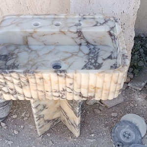 May include: A rectangular marble sink with a fluted exterior and a patterned interior. The sink has two faucet holes and a central drain. It is supported by a marble base. A round marble sink is also visible.
