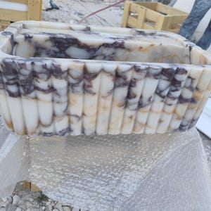 Könnte beinhalten: Rechteckiges Waschbecken aus Marmor mit gewelltem Rand. Das Waschbecken hat eine cremefarbene Basis mit dunkelvioletten und braunen Adern. Die Außenseite hat vertikale, geriffelte Details. Ein einzigartiges Badezimmer-Accessoire.