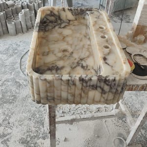 May include: A rectangular marble sink with a cream and brown veined pattern. The sink has a fluted exterior and three circular holes on the right side. It is set on a wooden stand in a workshop setting.