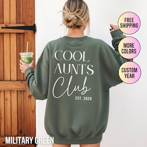 Könnte beinhalten: Militärgrünes Sweatshirt mit dem weißen Text "COOL AUNTS Club EST. 2025". Das Sweatshirt hat einen Rundhalsausschnitt. Das Bild zeigt auch ein Getränk in einem transparenten Plastikbecher.