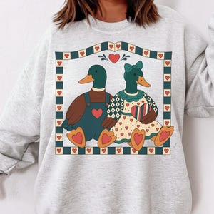 May include: A light gray sweatshirt featuring a whimsical illustration of two ducks in love. The ducks are inside a decorative frame with hearts. One duck wears overalls, the other a dress, both with heart accents.