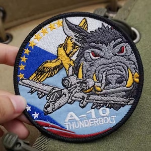 Puede incluir: Parche redondo bordado con una ilustración detallada de un avión A-10 Thunderbolt y un jabalí gruñón. El parche incluye el texto "A-10 THUNDERBOLT" y está bordeado por un borde negro. El diseño tiene una combinación de colores azul, blanco y dorado.