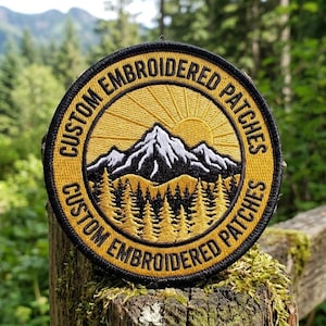 Op de afbeelding: Ronde, geborduurde patch met een berg- en zonontwerp in geel en zwart. De tekst "CUSTOM EMBROIDERED PATCHES" is in een cirkel rond de bergafbeelding geborduurd. De patch wordt buiten gepresenteerd, tegen een achtergrond van bomen.
