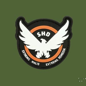 Op de afbeelding: Een zwart, wit en oranje embleem met een gestileerde adelaar met uitgespreide vleugels. De tekst "SHD" staat boven de adelaar en "EXTREMIS MALIS, EXTREMA REMEDIA" staat rond de rand.