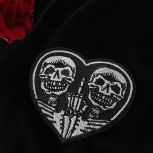以下が含まれることがあります： ハート型の2つの骸骨が刺繍された黒と白のパッチ。1つの骸骨が中指を立てています。