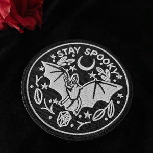 以下が含まれることがあります： コウモリ、三日月、星、葉が刺繍された黒と白のパッチ。パッチの縁には「Stay Spooky」という文字が書かれています。