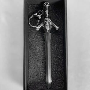Op de afbeelding: Een zilverkleurige zwaardvormige sleutelhanger met een skeletontwerp, gepresenteerd in een zwarte doos. De sleutelhanger heeft een gedetailleerde skeletkop en armen, met een ring om sleutels aan te bevestigen. Het zwaard heeft een punt.