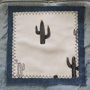 May include: A square fabric patch with a denim border and a white center. The center features a black and white cactus design. The patch is sewn onto a light blue denim background with white stitching around the border.