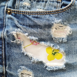 May include: Blue denim shorts featuring distressed areas with frayed edges and holes. A white fabric patch with a yellow and pink floral print is visible underneath the largest distressed section. The shorts include copper-colored metal rivets.