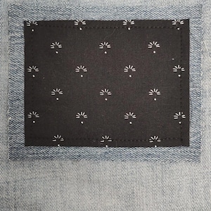 May include: Close-up of light blue denim jeans with a black patch. The patch is rectangular and features a repeating white floral pattern. The patch is stitched onto the jeans.
