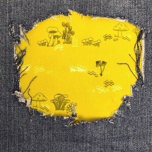 May include: A pair of blue denim jeans featuring a large, distressed, frayed-edge hole that reveals a bright yellow fabric patch underneath. The yellow patch is decorated with a repeating pattern of various black and white line-art mushrooms.