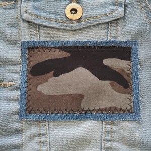 May include: Close-up of a denim jacket with a rectangular camouflage patch. The patch features a brown, gray, and black pattern, framed by a blue denim border and stitched with dark thread. A button is visible at the top.