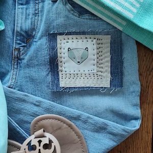 May include: Light blue denim jeans with a fox patch. The patch is square with a white background and a gray fox illustration. A pair of beige sandals and a turquoise and white striped towel are also in the image.