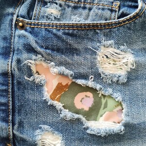 May include: Blue denim shorts featuring distressed areas with frayed edges and exposed patches of fabric underneath. The patches display a floral pattern in shades of olive green, light pink, and brown, accented with metallic copper-colored rivets.