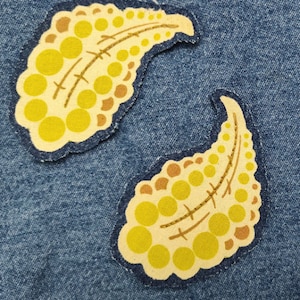 May include: Two yellow and brown paisley-shaped fabric patches with a stitched design of circles and lines. The patches are on a blue denim background.