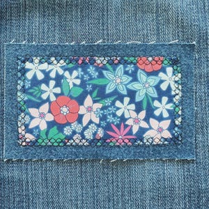 May include: Close-up of a denim patch with a floral design. The patch features a dark blue background with white, pink, and light blue flowers. The patch is stitched onto a denim background with a black thread.