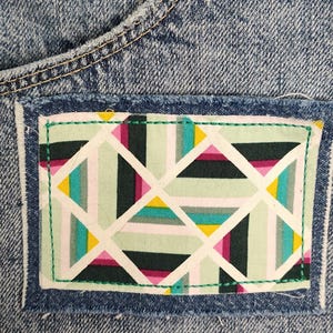 May include: A blue denim fabric garment featuring a rectangular patch with a geometric pattern. The patch displays intersecting white lines forming triangles and rectangles in shades of mint green, dark green, magenta, yellow, and teal.