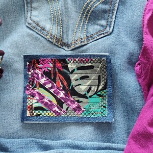 May include: A close-up of a pair of blue jeans with a colorful fabric patch sewn over a hole in the pocket. The patch features a geometric pattern with pink, purple, and green accents.