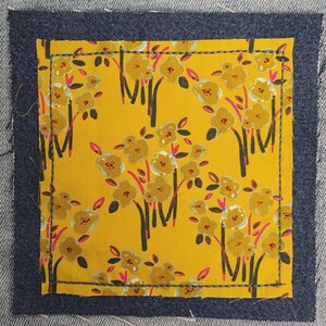 May include: Close-up of a denim patch with a yellow floral pattern. The patch is rectangular with a dark blue border and is sewn onto a light blue denim fabric. The floral design features yellow flowers and black stems.