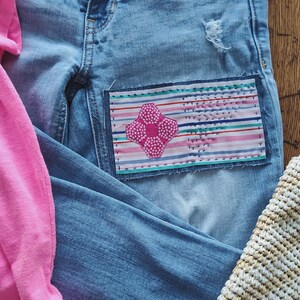 May include: Light blue denim jeans with a distressed knee and a decorative patch. The patch features a pink and white floral design on a striped background. A pink shirt, sandals, and a woven bag are also visible.