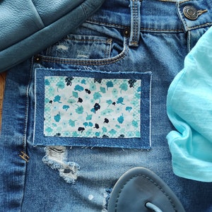 May include: Blue denim jeans with a small floral patch sewn on the front pocket. The jeans have a distressed look with a small hole in the fabric. The jeans are paired with a light blue t-shirt and blue leather sandals.