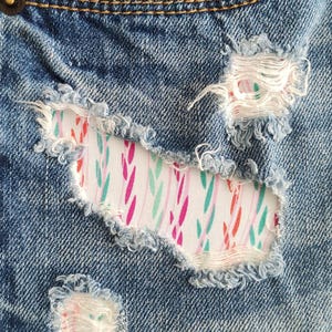 May include: Blue denim fabric featuring distressed, frayed holes that reveal a white fabric patch underneath. The patch is decorated with a repeating pattern of small, vertical, leaf-like shapes in shades of bright pink, teal, and orange.