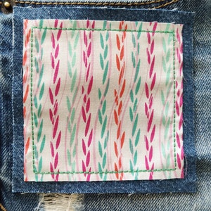 May include: A close-up of a denim fabric with a small patch of fabric sewn on. The patch has a white background with a repeating pattern of pink, green, and orange leaves.