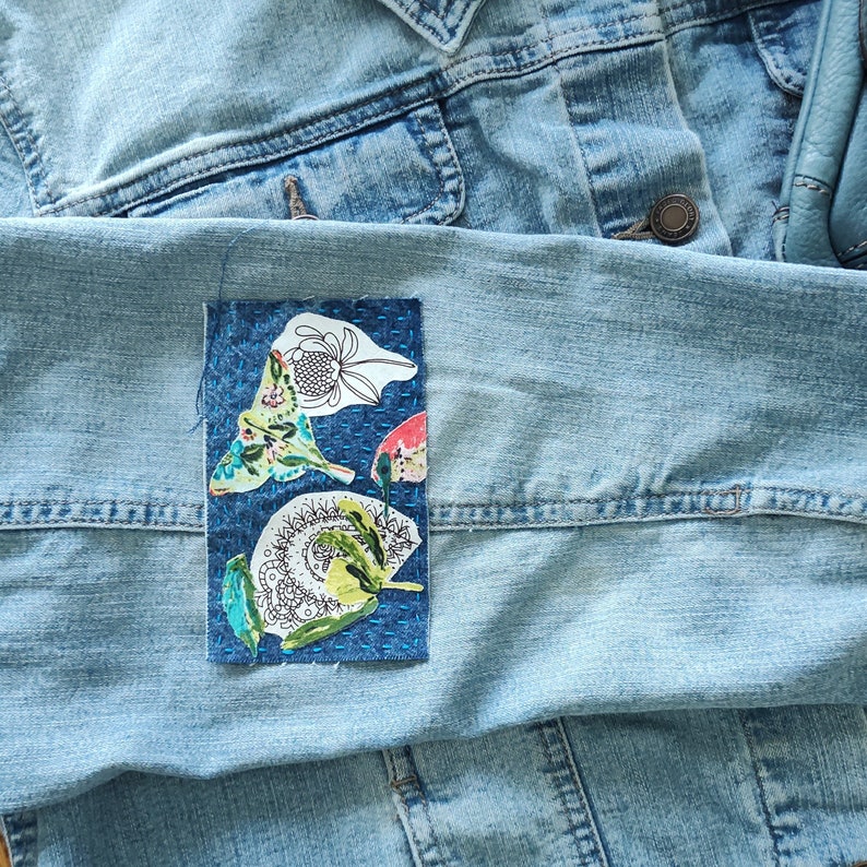 May include: A blue denim jacket with a colorful fabric patch sewn onto the left side. The patch features a floral design with butterflies and a swirl pattern.