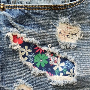 May include: Blue denim jeans with a large floral patch sewn over a ripped area. The patch is made of a blue fabric with white, pink, and green flowers.