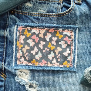 May include: Blue denim jeans featuring a rectangular fabric patch with a dark gray background and a repeating pattern of small yellow, pink, and white butterflies. The patch is stitched with light pink thread and has frayed edges.