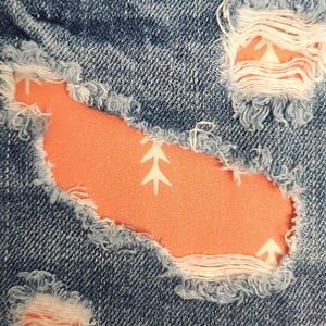 May include: Blue denim fabric featuring distressed, frayed-edge cutouts that reveal a bright orange fabric backing. The orange fabric is decorated with a repeating white pattern of small, stylized arrow-like shapes pointing upwards.