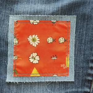May include: Close-up of a denim patch sewn onto blue jeans. The patch is a square of orange fabric with a floral and ladybug pattern. The patch is bordered by a light blue denim square and stitched with gray thread.