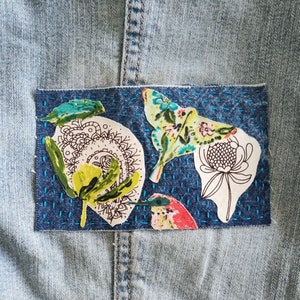 May include: A blue denim jacket with a patch of fabric featuring a variety of colorful floral and insect designs. The patch is stitched onto the jacket with blue thread.