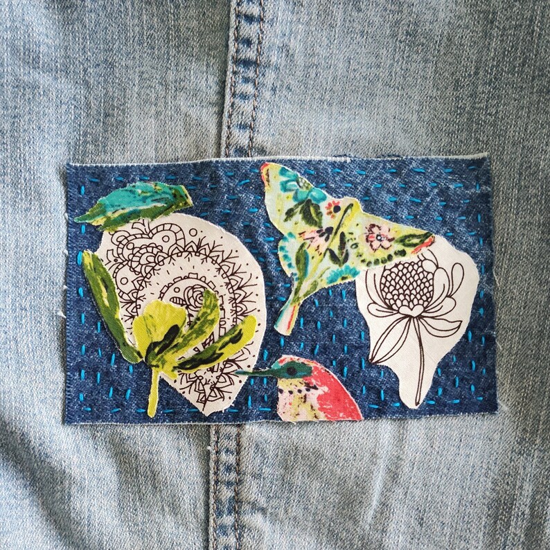 May include: A blue denim jacket with a patch of fabric featuring a variety of colorful floral and insect designs. The patch is stitched onto the jacket with blue thread.
