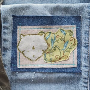 May include: Close-up of a denim patch with a light blue border. Inside, a light pink square features a white cat face and a green and blue floral design. The patch is sewn onto a light blue denim fabric.