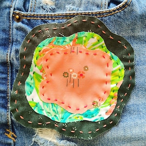 May include: A dark green fabric patch with a pink, green, and white floral design sewn onto a blue denim background.