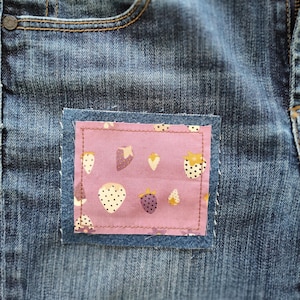 May include: A pair of blue denim jeans featuring a rectangular fabric patch sewn onto the front. The patch is light purple and decorated with a repeating pattern of small, stylized strawberries in white, yellow, and dark purple colors.