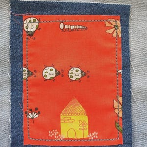 May include: A denim patch with a stitched orange fabric square featuring a whimsical design. The orange fabric displays ladybugs, a house, flowers, and a caterpillar. The patch is sewn onto a denim background.