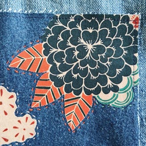 May include: A light blue denim fabric featuring a rectangular patch with a dark teal floral pattern, orange and green striped leaves, and a light pink cloud-like shape with red dots, all secured with visible white and orange decorative stitching.