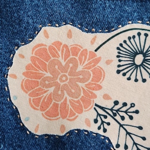 May include: A blue denim fabric garment featuring a beige fabric patch stitched onto the surface. The patch displays a printed design of a large orange flower, a dark green dandelion-like seed head, and a dark green leafy branch motif.