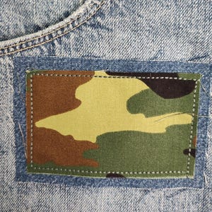 May include: A light blue acid-wash denim fabric featuring a rectangular patch sewn onto the surface. The patch displays a camouflage pattern in shades of olive green, brown, and black. The denim includes a metal rivet and frayed edges.