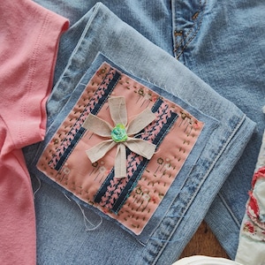 May include: Light blue denim jeans with a pink patch featuring a flower design. A pink t-shirt and floral patterned shoes are also visible. The patch is square, with a floral design and a ribbon flower in the center.