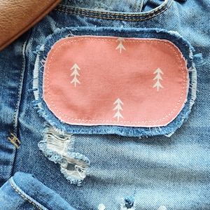 May include: A pair of blue denim jeans with a pink fabric patch sewn on the front. The patch has a white outline and a repeating pattern of white stylized trees.