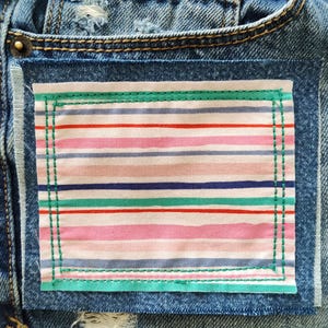 May include: A pair of blue denim jeans featuring a rectangular fabric patch sewn onto the front. The patch displays a horizontal pattern of thin stripes in shades of pink, red, blue, and green, secured with visible green stitching around the edges.